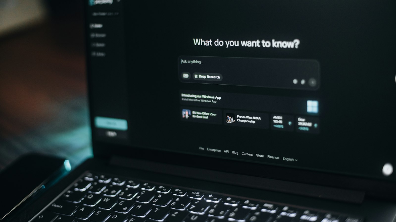A laptop screen glowing in a dark room shows an AI search interface reading ‘What do you want to know?’ above an ‘Ask anything…’ prompt bar